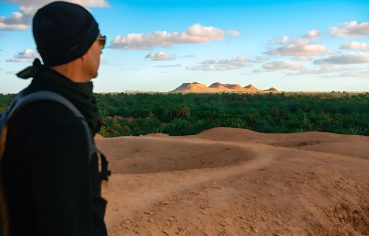 A man in a black beanie and sunglasses looking out over the dense palm groves of the Siwa Oasis from a sandy viewpoint, with rounded, low mountains visible in the distance under a blue sky.