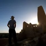 A silhouette of a Man Looking At The Temple Of The Oracle Amun in Siwa Oasis at sunset, with the sun setting directly behind the tall, ruined mud-brick structure.