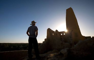 A silhouette of a Man Looking At The Temple Of The Oracle Amun in Siwa Oasis at sunset, with the sun setting directly behind the tall, ruined mud-brick structure.