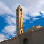 The tall, narrow bell tower of Saint Pishoy Monastery in Wadi El Natrun against a bright blue, cloudy sky.