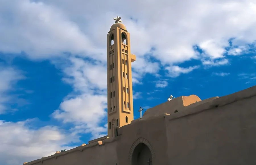 The tall, narrow bell tower of Saint Pishoy Monastery in Wadi El Natrun against a bright blue, cloudy sky.