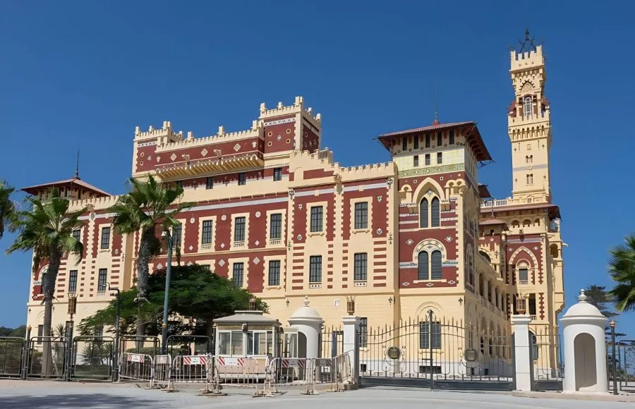A colorful, wide view of Montaza Palace Outside in Alexandria, showing the ornate yellow and red historical structure, a tall clock tower, and palm trees in the foreground against a clear blue sky.