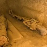 Two ancient mummies wrapped and partially unwrapped, lying in a carved niche inside a tomb on the Mountain of the Dead in Siwa Oasis, with the tomb's interior illuminated in a warm glow.