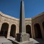 A towering black Obelisk in the German cemetery at El Alamein, surrounded by an arched, curved stone courtyard and a clear blue sky above.