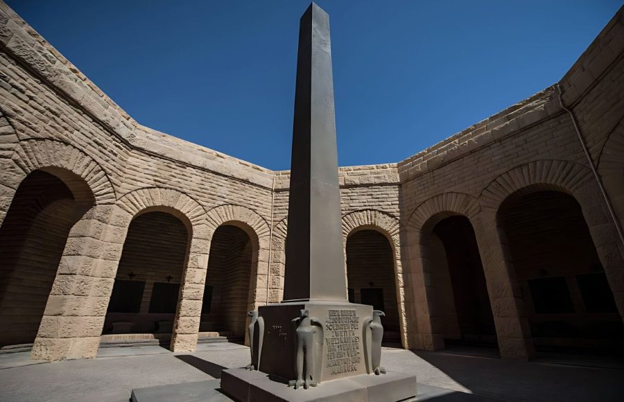 A towering black Obelisk in the German cemetery at El Alamein, surrounded by an arched, curved stone courtyard and a clear blue sky above.