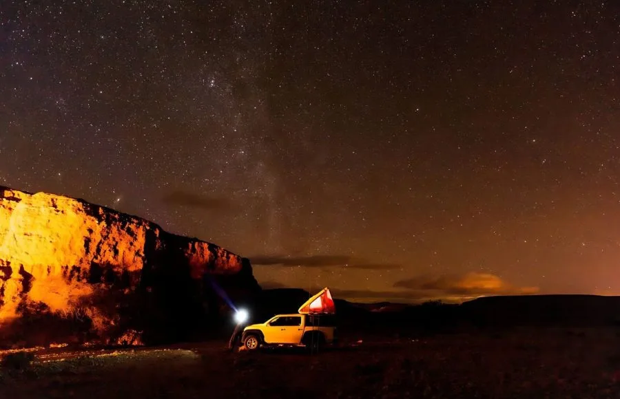Off-road car with a rooftop tent illuminated at a desert camp at night under a starry sky.