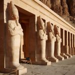 A row of large, standing Osiride statues, depicting the pharaoh, lining the colonnade at the Osiris Statues At Temple Of Hatshepsut in Luxor, Egypt.