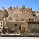 A bright, clear view of the magnificent Outside View Shali Fortress Old Siwa, showing the large, crumbling, mud-brick ruins towering over shops and bicycles in the foreground.