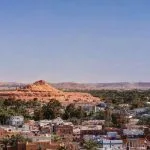 A wide Panorama Of The City Of Shali in Siwa Oasis, showing the modern town and lush palm groves in the foreground, with the high, rocky, ruined mountain of Old Shali rising in the middle ground.