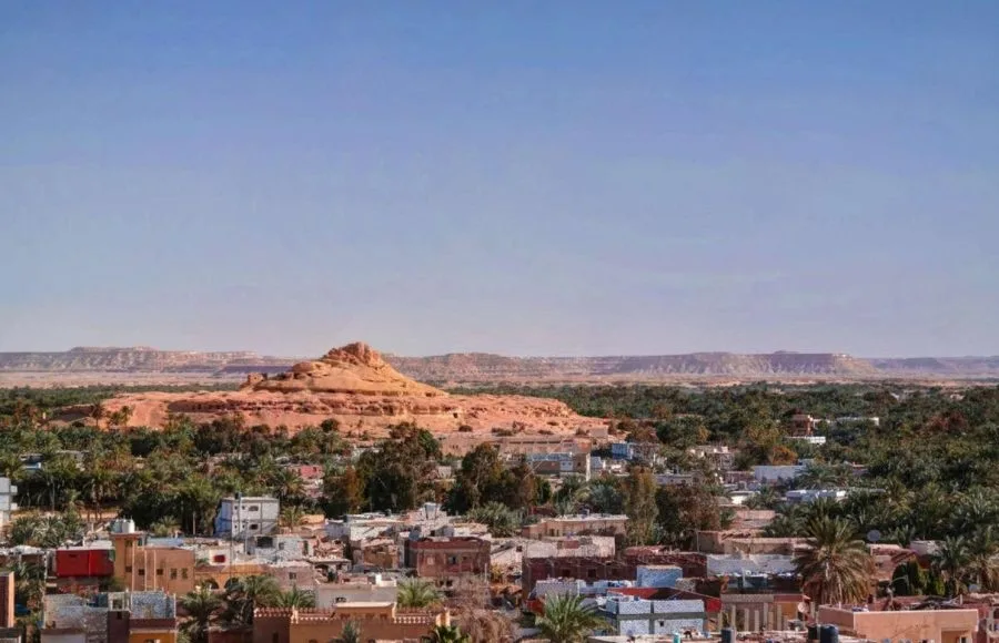 A wide Panorama Of The City Of Shali in Siwa Oasis, showing the modern town and lush palm groves in the foreground, with the high, rocky, ruined mountain of Old Shali rising in the middle ground.
