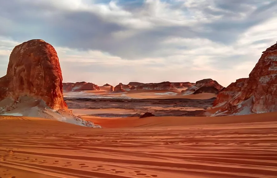 A wide Panorama of El Agabat Valley in the White Desert, showing large reddish and white rock formations against a sky with dramatic clouds.