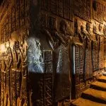 Dark, dramatic view of the richly carved Reliefs At Interior Temple Of Hathor In Dendera, showing hieroglyphs and figures illuminated by warm light on a sloping passage.