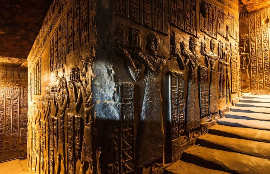 Dark, dramatic view of the richly carved Reliefs At Interior Temple Of Hathor In Dendera, showing hieroglyphs and figures illuminated by warm light on a sloping passage.