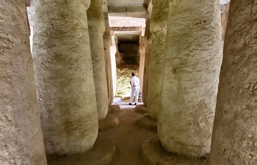 Rock-cut columns lining a passage, with a lone figure at the brightly lit end of a burial chamber at Amarna.