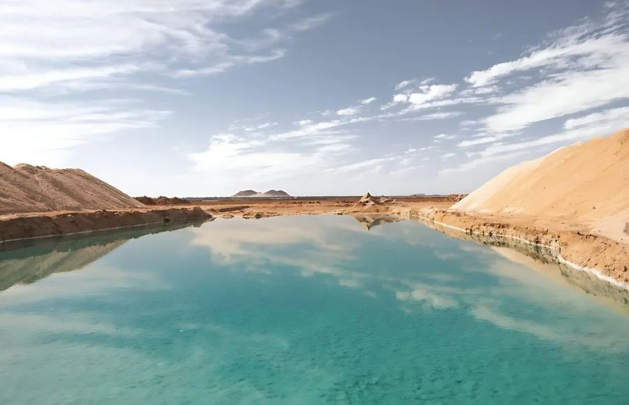 Wide panoramic view of a bright turquoise Salt Lake of Siwa, reflecting the sky and clouds, with banks of white and tan salt deposits surrounding the water.