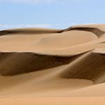 A close-up view of the majestic, layered Sea Dunes Of The Great Sand Sea, Egypt, showing steep slopes of golden and brown sand under a light blue sky with wispy clouds.