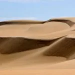 A close-up view of the majestic, layered Sea Dunes Of The Great Sand Sea, Egypt, showing steep slopes of golden and brown sand under a light blue sky with wispy clouds.