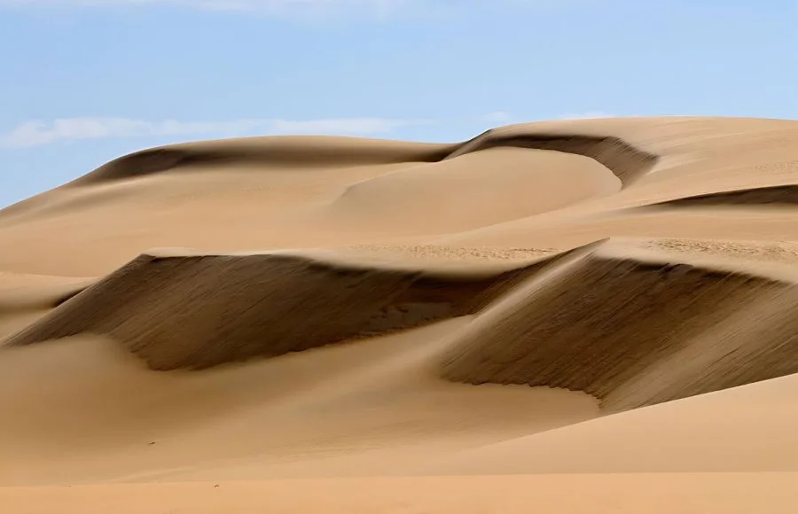 A close-up view of the majestic, layered Sea Dunes Of The Great Sand Sea, Egypt, showing steep slopes of golden and brown sand under a light blue sky with wispy clouds.