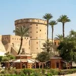 A Sideview Of Citadel Salah Eldin in Cairo, showing the massive, round-towered fortress walls towering over a lush green garden area with palm trees.