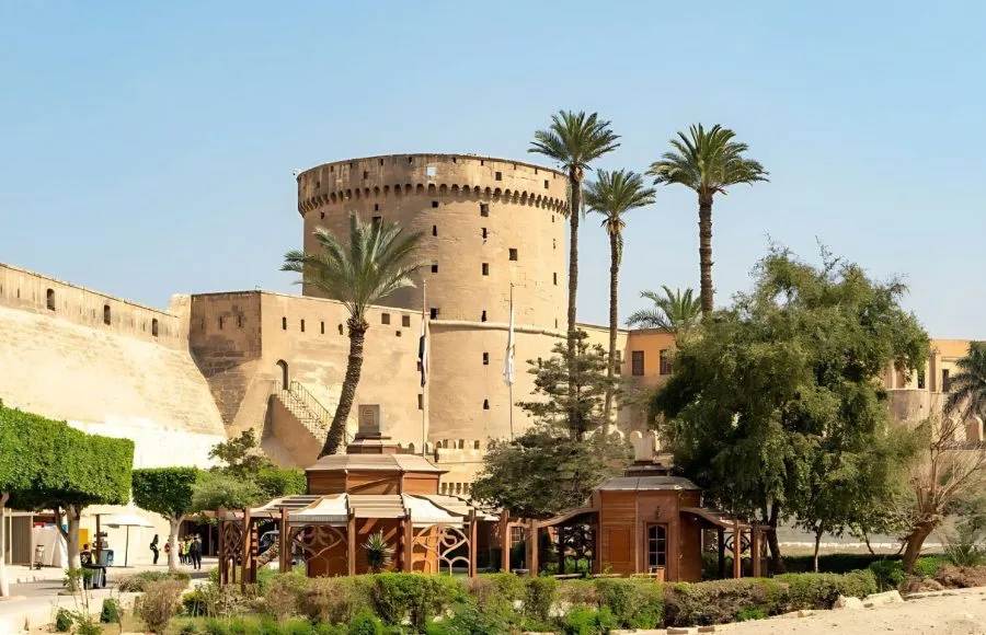 A Sideview Of Citadel Salah Eldin in Cairo, showing the massive, round-towered fortress walls towering over a lush green garden area with palm trees.