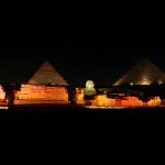 Three Giza Pyramids and the Sphinx illuminated by warm orange floodlights at midnight.