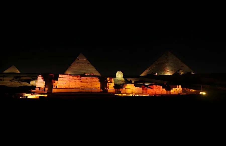 Three Giza Pyramids and the Sphinx illuminated by warm orange floodlights at midnight.