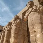 Colossal Stone Figures RamsesII As Osiris In The Karnak Temple Complex, viewed from a low angle against a blue sky, showcasing the ancient carvings.
