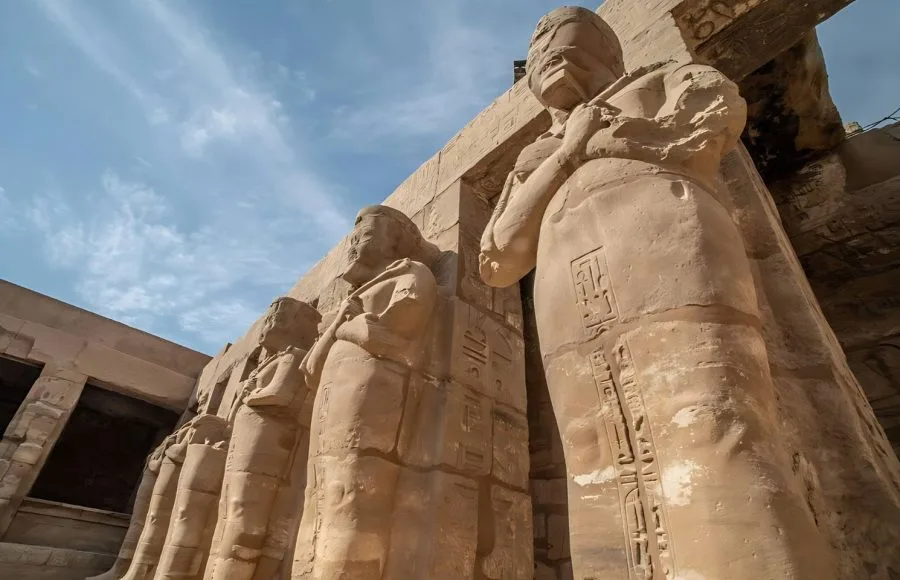 Colossal Stone Figures RamsesII As Osiris In The Karnak Temple Complex, viewed from a low angle against a blue sky, showcasing the ancient carvings.