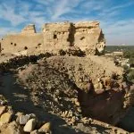 The sunlit, crumbling stone ruins of the Temple of the Aghurmi in Siwa Oasis, perched on a rocky mound overlooking a lush grove of palm trees and the oasis landscape.