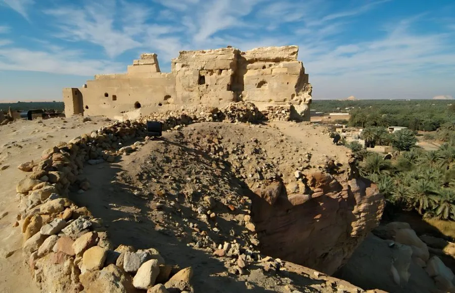 The sunlit, crumbling stone ruins of the Temple of the Aghurmi in Siwa Oasis, perched on a rocky mound overlooking a lush grove of palm trees and the oasis landscape.