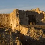 The sunlit, weathered stone ruins of the Temple of the Oracle in Siwa Oasis, Egypt, featuring rough, ancient walls and stepped rubble under a clear blue sky.