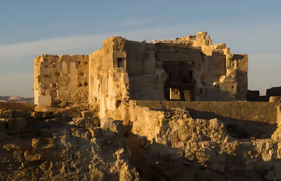 The sunlit, weathered stone ruins of the Temple of the Oracle in Siwa Oasis, Egypt, featuring rough, ancient walls and stepped rubble under a clear blue sky.
