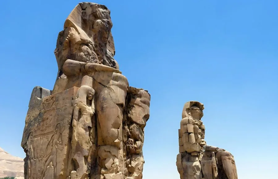 The towering Colossi of Memnon, two massive stone statues of Pharaoh Amenhotep III, under a clear blue Egyptian sky.