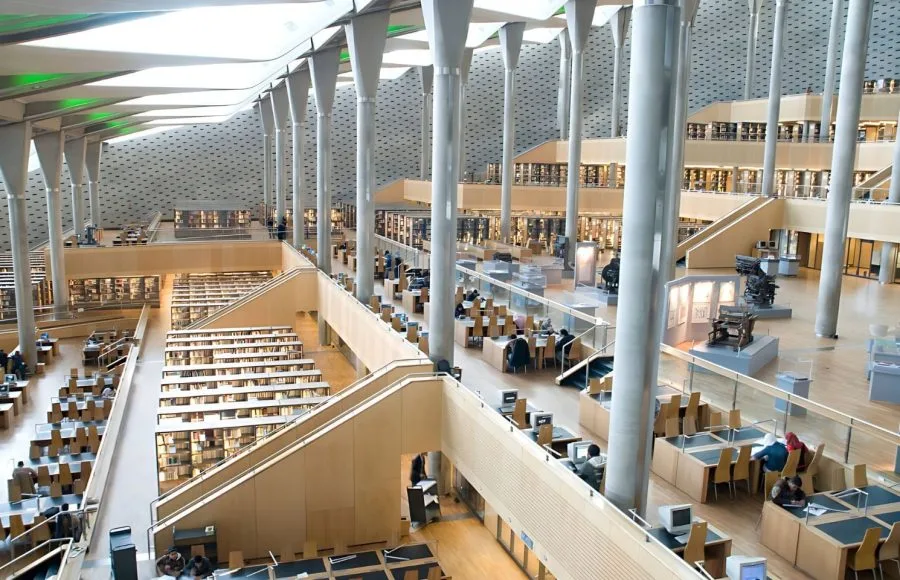 A multi-level, high-angle view of The Interior Rotunda Of Alexandrina library (Bibliotheca Alexandrina), featuring modern architecture, light gray columns, reading desks, and vast rows of bookshelves.