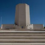 The large, modern, tiered, white stone monument of The Italian Memorial in El Alamein, featuring a tall central tower and wide staircase leading up to the entrance against a bright blue sky.