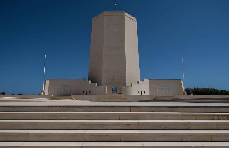 The large, modern, tiered, white stone monument of The Italian Memorial in El Alamein, featuring a tall central tower and wide staircase leading up to the entrance against a bright blue sky.
