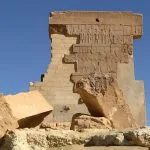 The large, freestanding, crumbling stone wall of The Temple of Umm Ubaydah in Siwa, Egypt, showing preserved carved reliefs on the facade and surrounded by massive fallen stone blocks under a clear blue sky.