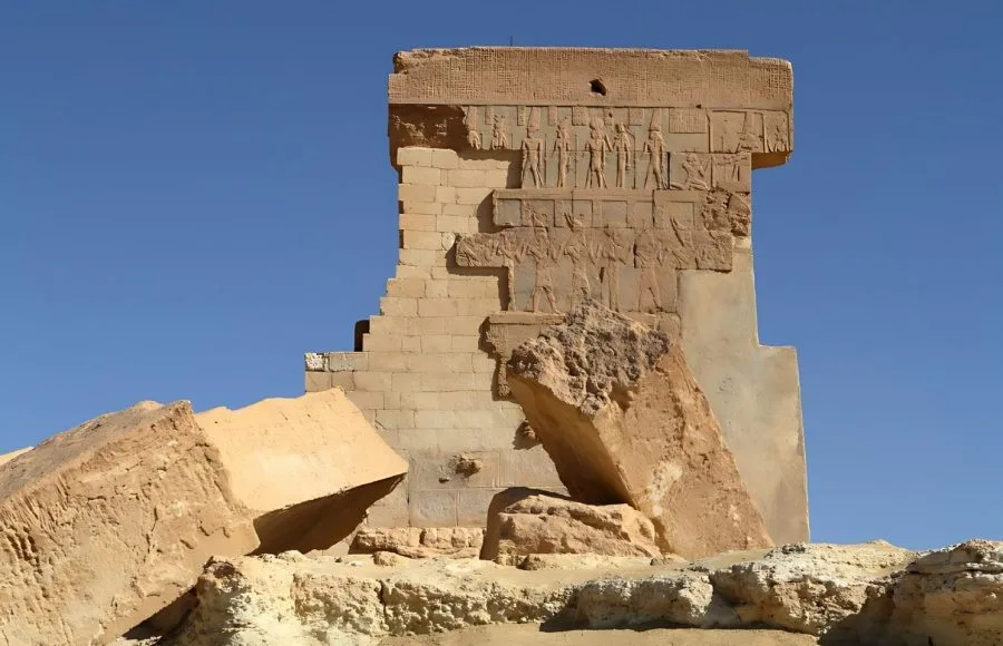 The large, freestanding, crumbling stone wall of The Temple of Umm Ubaydah in Siwa, Egypt, showing preserved carved reliefs on the facade and surrounded by massive fallen stone blocks under a clear blue sky.