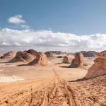 A panoramic view of the Valley of Agabat in the White Desert, Egypt, featuring soft white and light brown wind-eroded rock formations contrasting against the sandy ground and a blue sky with clouds.