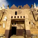The highly decorated, mud-brick facade of the Badr Museum in Farafra Oasis, Egypt, featuring relief carvings of camels, antelopes, palm trees, and Egyptian life under a bright blue sky.