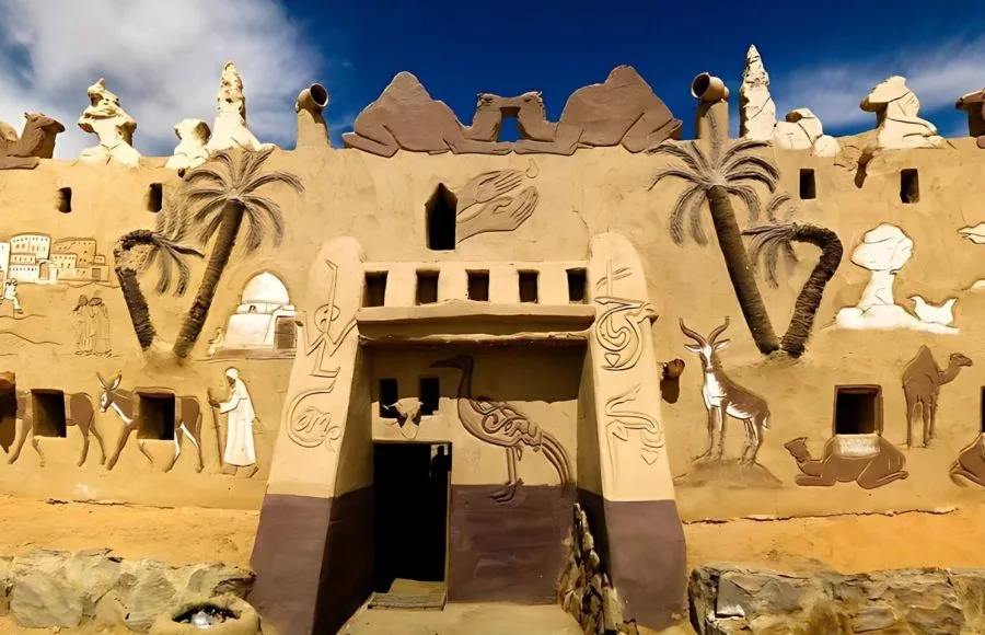 The highly decorated, mud-brick facade of the Badr Museum in Farafra Oasis, Egypt, featuring relief carvings of camels, antelopes, palm trees, and Egyptian life under a bright blue sky.