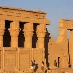A Tourist Couple stands in front of the colossal, ornate columns and the preserved wall carvings of the colonnade of the temple of Hathor at Dendera, bathed in golden light.
