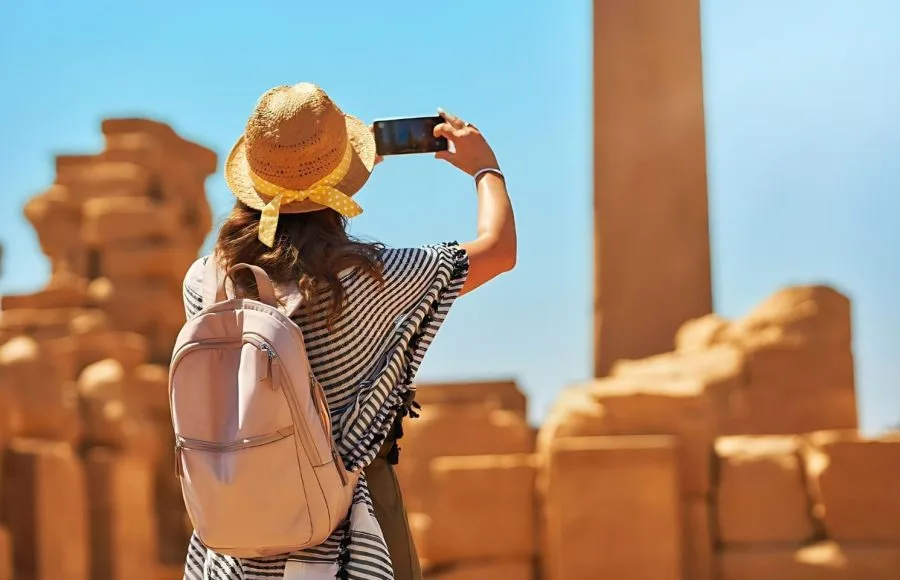 A female Tourist wearing a straw hat and backpack, taking a photo with a smartphone of the massive sandstone columns and ruins in the Karnak Temple complex in Luxor, Egypt.
