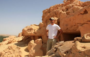 A male tourist in a white shirt and khaki shorts stands in front of a carved tomb entrance in the yellow, sun-drenched sandstone of the Mountain of the Dead in Siwa Oasis.
