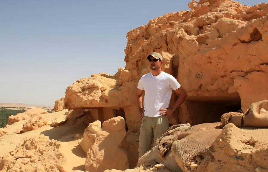 A male tourist in a white shirt and khaki shorts stands in front of a carved tomb entrance in the yellow, sun-drenched sandstone of the Mountain of the Dead in Siwa Oasis.