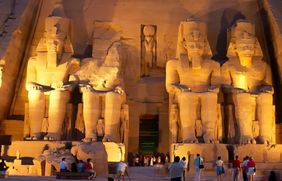 Lit-up night view of the Great Temple of Ramesses II at Abu Simbel, Egypt, featuring colossal seated statues and tourists.