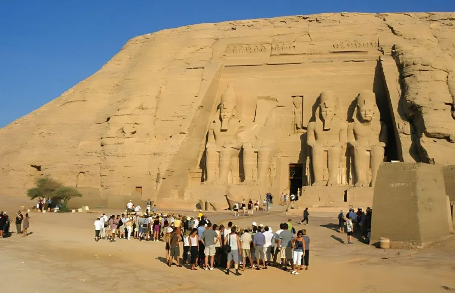 A large group of Tourists standing in front of the Great Temple of Ramses II at Abu Simbel, viewing the four massive seated statues carved into the cliff face.