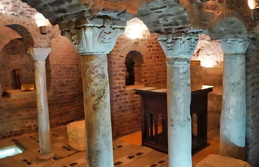 The dimly lit, ancient stone arches and pillars of the underground View Of The Crypt Abu Sergius Church, showing rough-hewn columns and simple steps leading down.