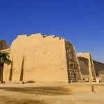 The colossal exterior walls of the View of the Medinet Habu Temple in Luxor, showing the side perspective of the ancient Egyptian structure against a clear blue sky.