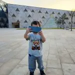 A Young Tourist Child stands outside the modern, geometric facade of The Grand Egyptian Museum (GEM) and is Taking Selfie with a smartphone.